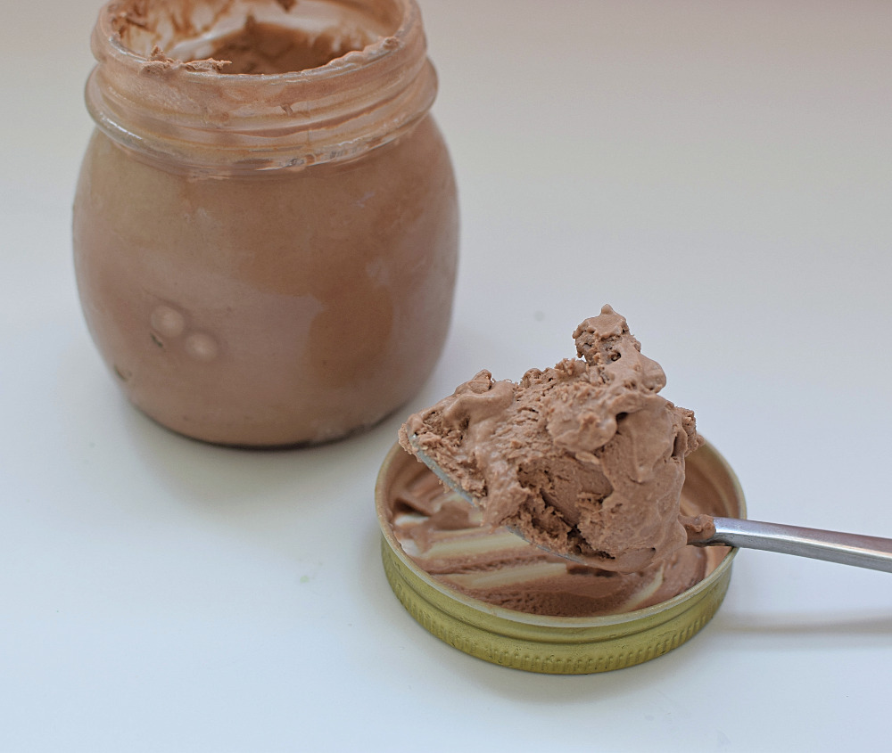 A jar of chocolate ice cream next to a spoon of ice cream.