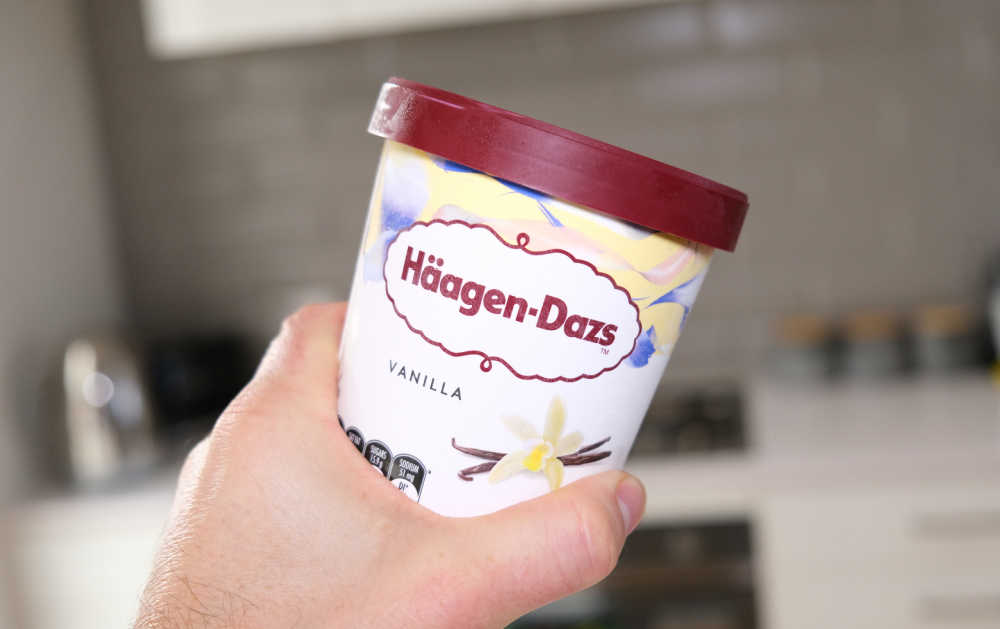 A hand holding an unopened pack of Haagen Dazs with the kitchen in the background, blurred.