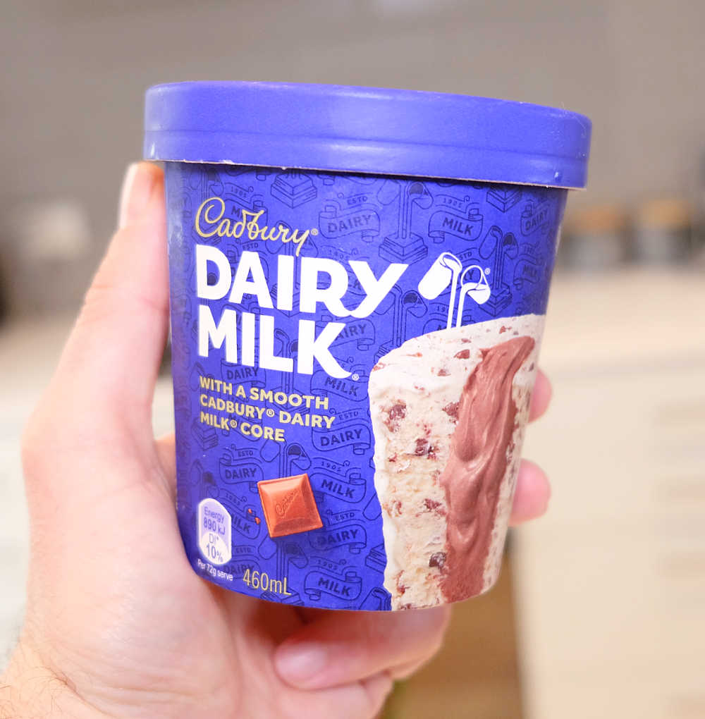 Closeup of a hand holding a tub of Dairy Milk with a blurred background.