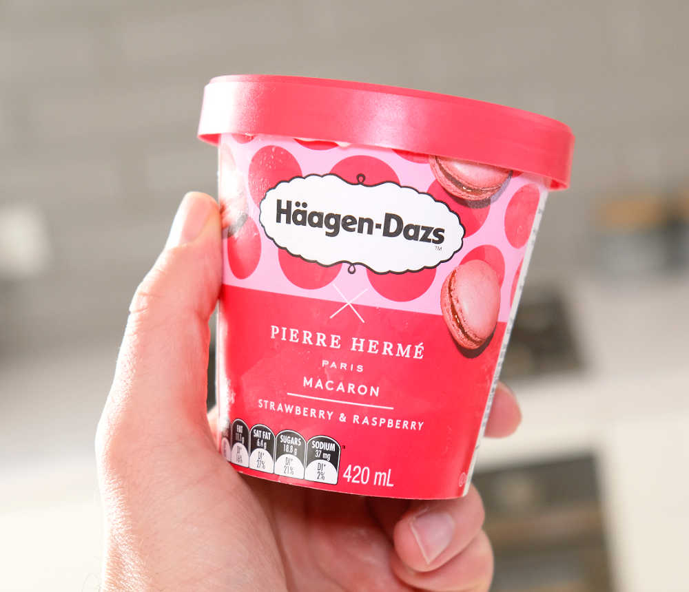 A hand holding a tub of Haagen-Dazs Pierre Herme Paris Macaron Strawberry and Raspberry Ice Cream.