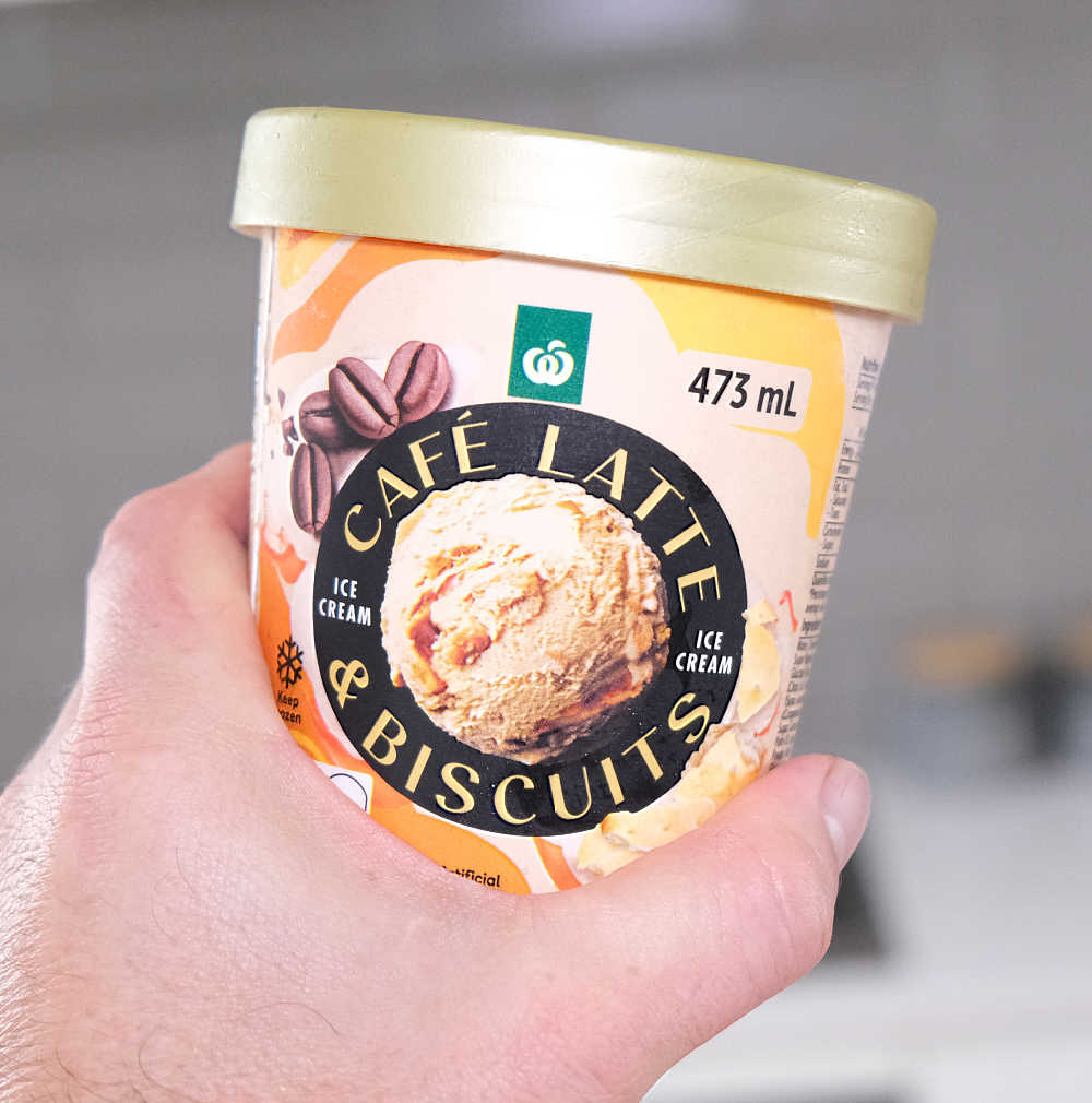 A hand holding a pack of Woolworths Cafe Latte & Biscuits Ice Cream with a blurred kitchen in the background.