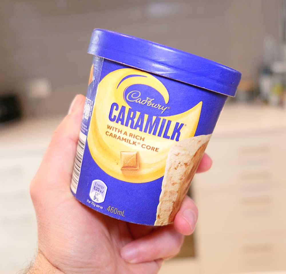 A hand holding a tub of Cadbury Caramilk with a blurred kitchen in the background.