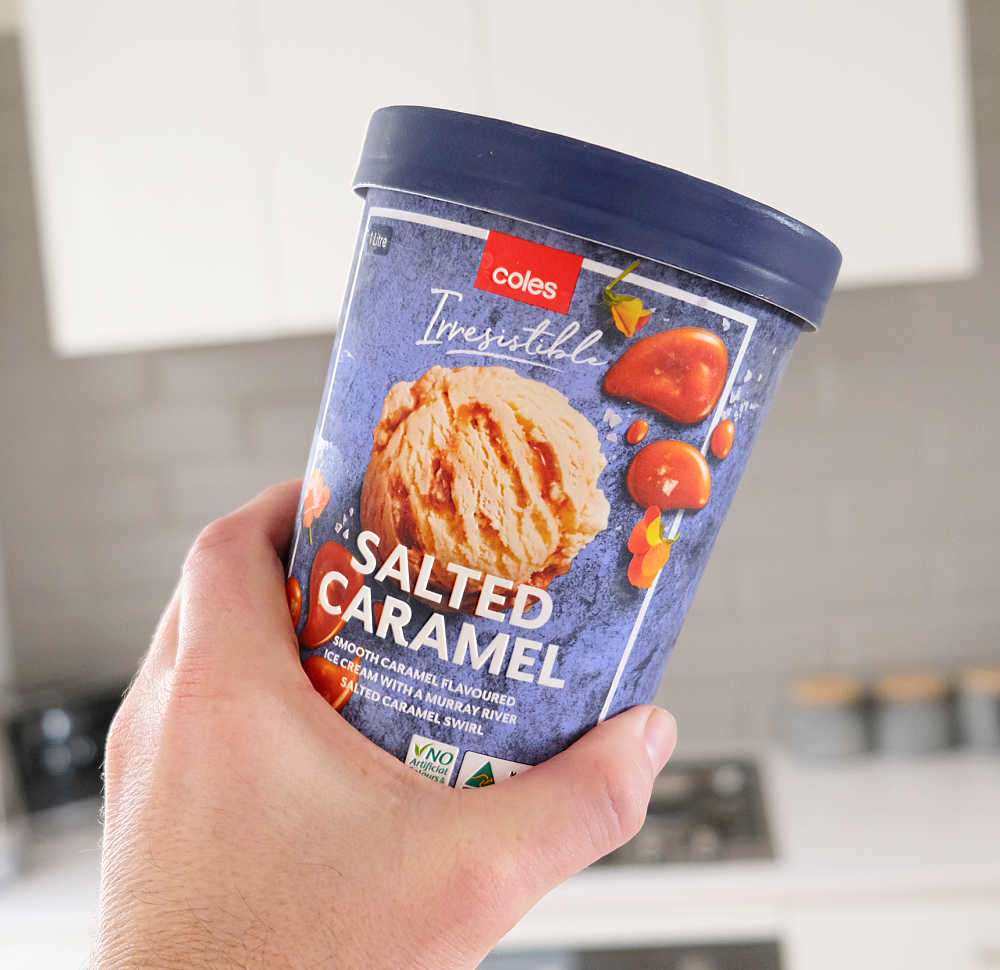 A hand holding a tub of Coles Salted Caramel ice cream with a blurred kitchen in the background.