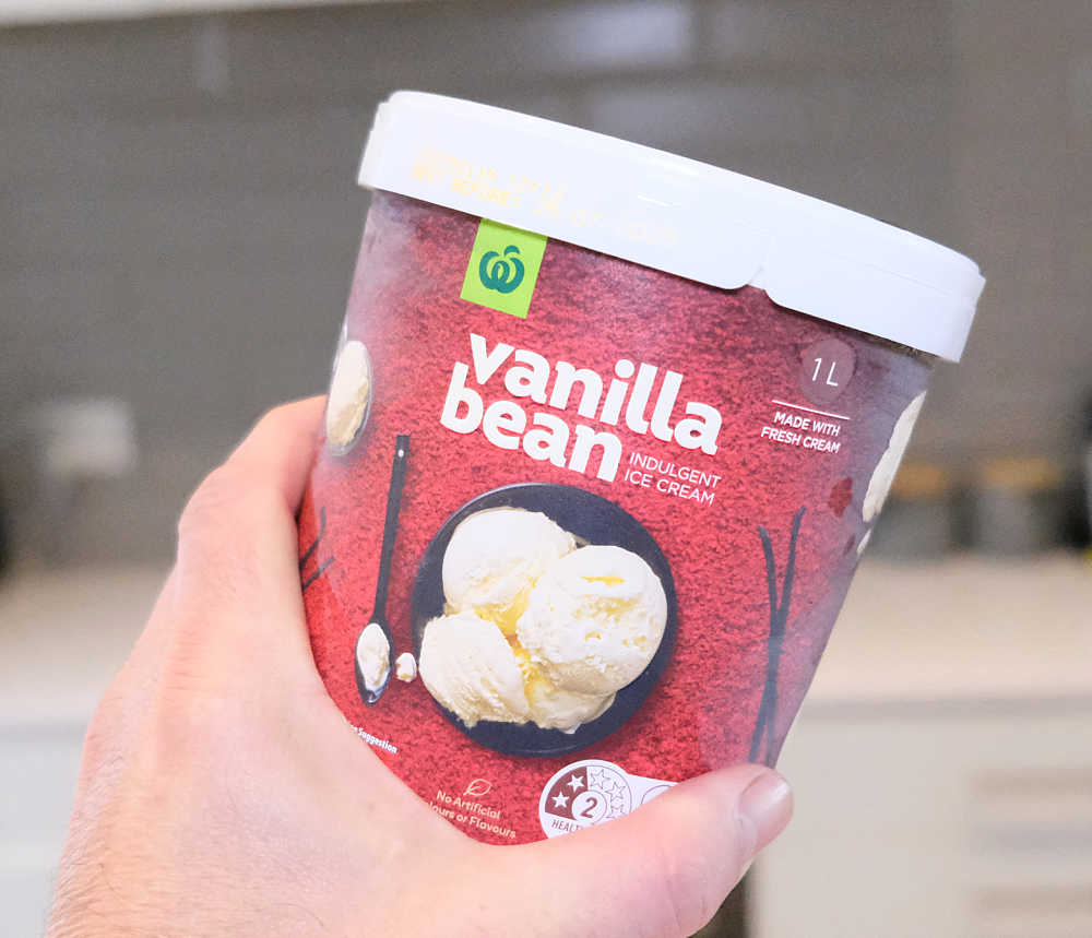 A hand holding a tub of Woolworths Vanilla Bean Ice Cream with a blurred kitchen in the background.