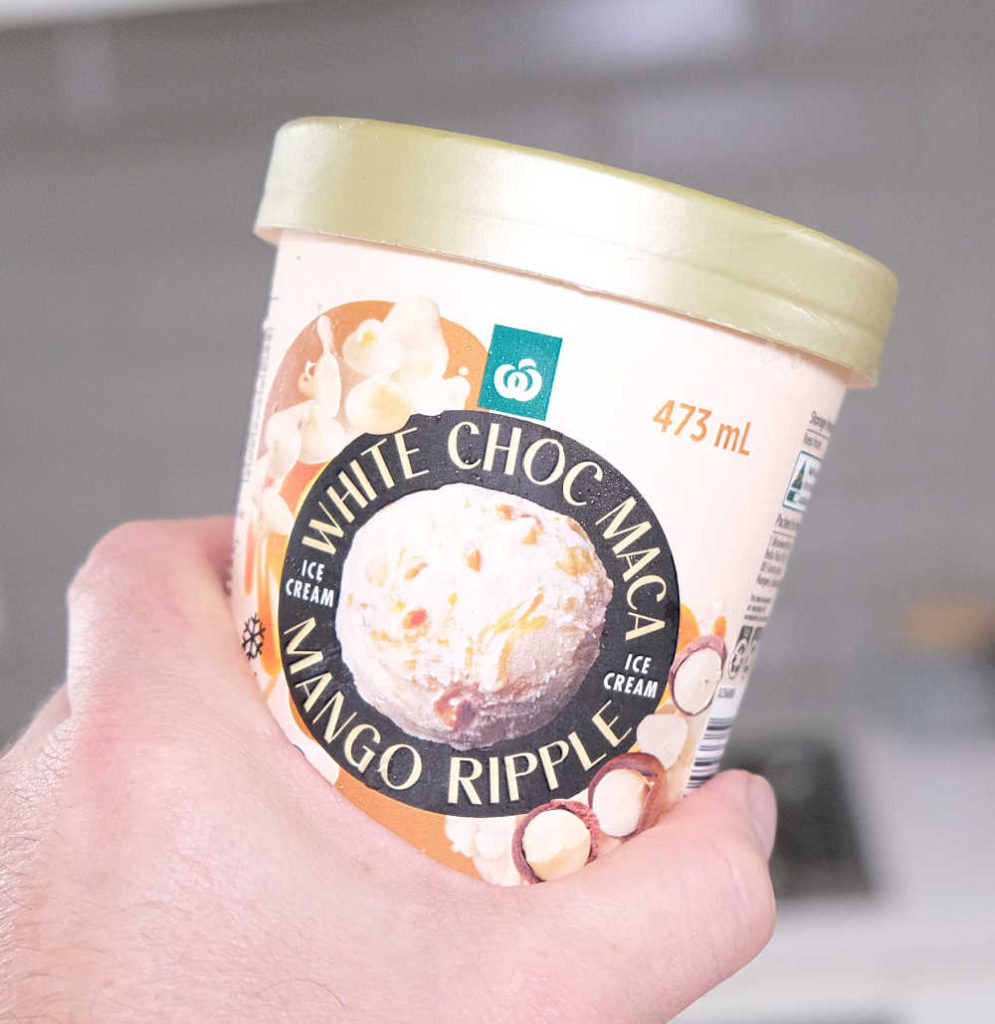 A hand holding a pint of Woolworths White Choc Maca Mango Ripple Ice Cream.