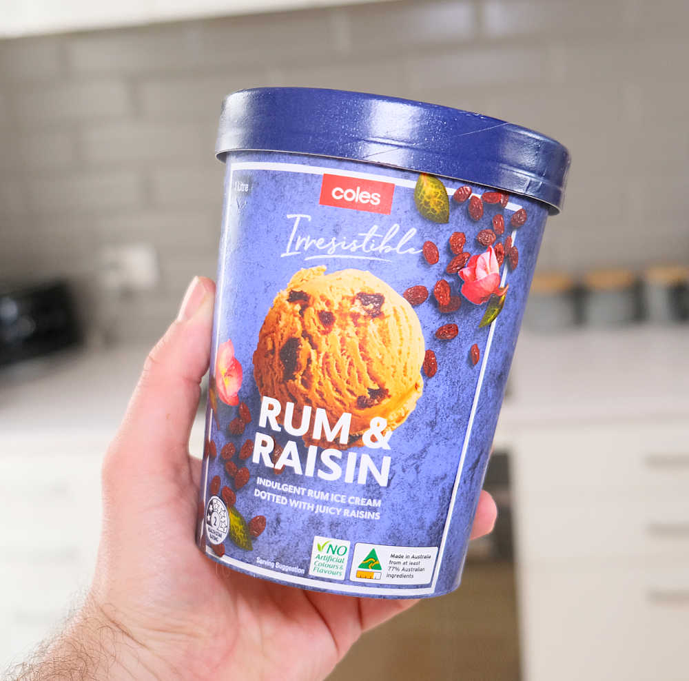 Holding a tub of Coles Rum and Raisin Ice Cream with a blurred kitchen in the background.