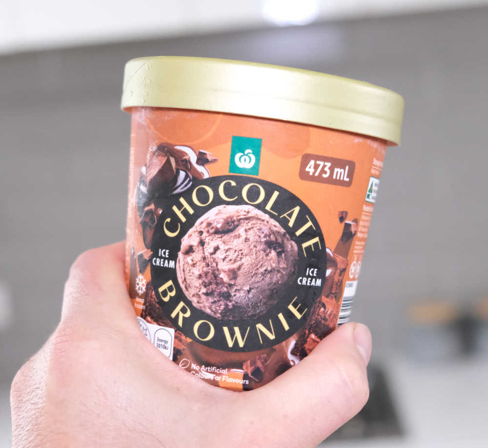 Holding a tub of Woolworths Chocolate Brownie Ice Cream with a blurred kitchen in the background.