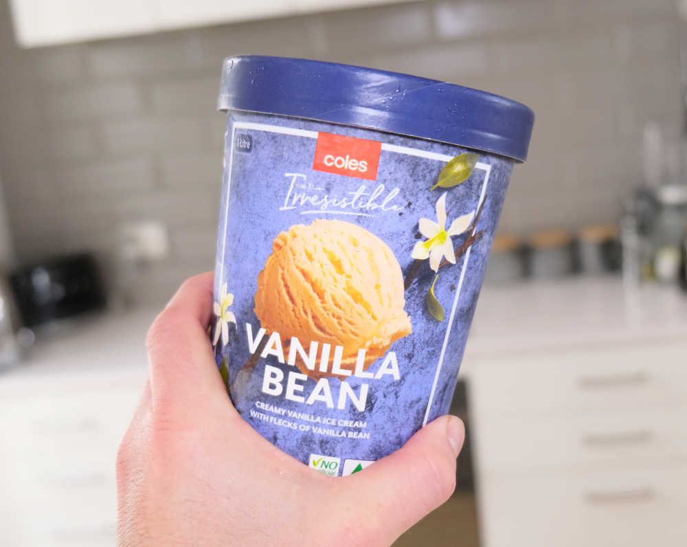 Holding a tub of Coles Irresistible Vanilla Bean Ice Cream with a blurry kitchen in the background.