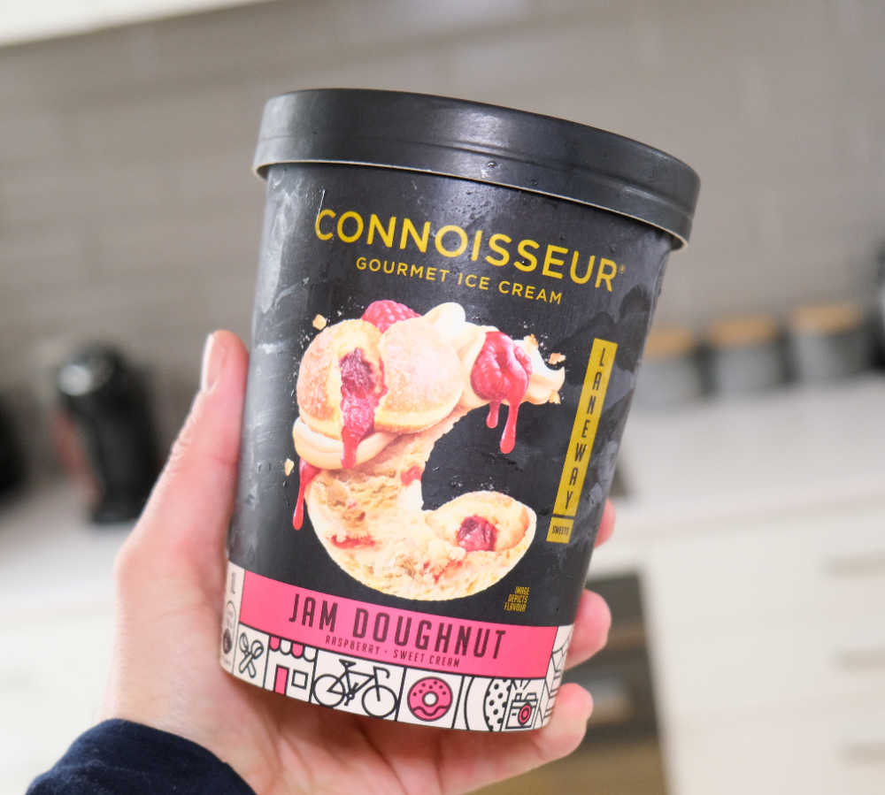 A hand holding a tub of Jam Doughnut ice cream with a blurred kitchen in the background.