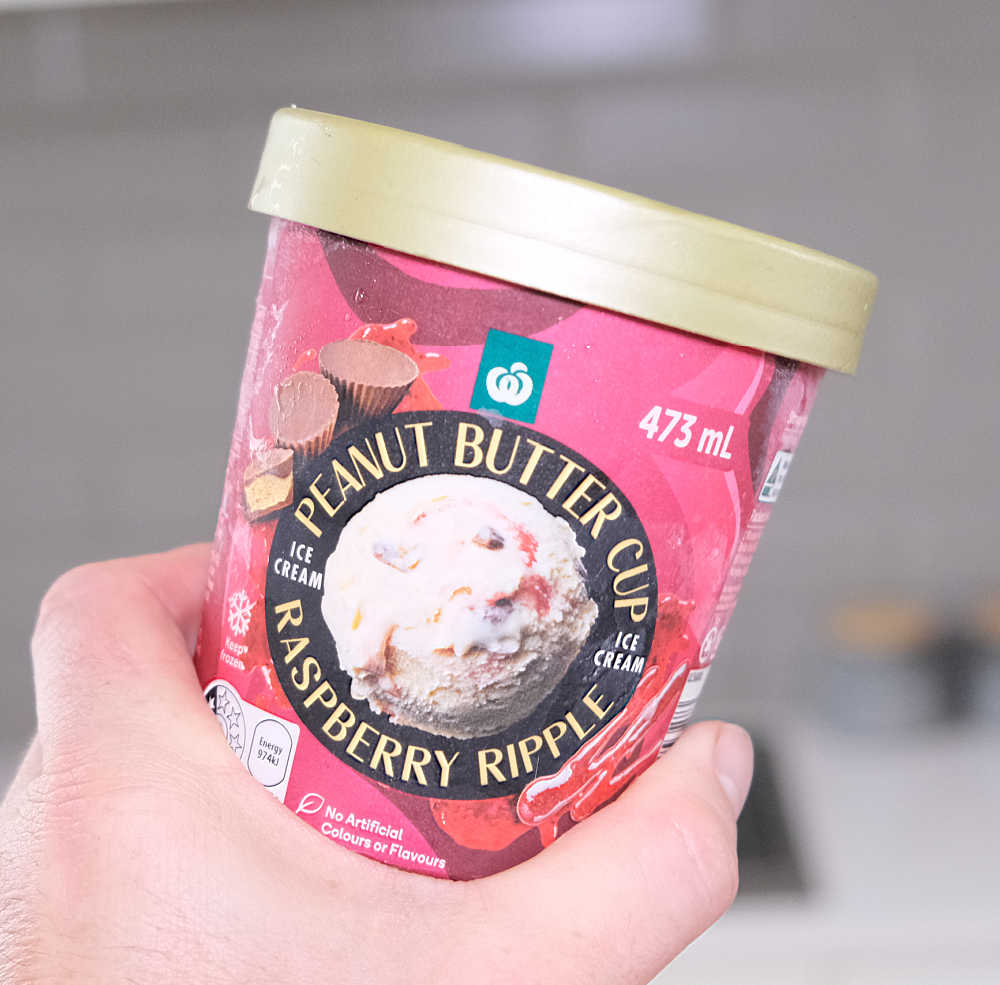 A hand hoding a container of Woolworths Peanut Butter Cup Raspberry Ripple Ice Cream 473ml.