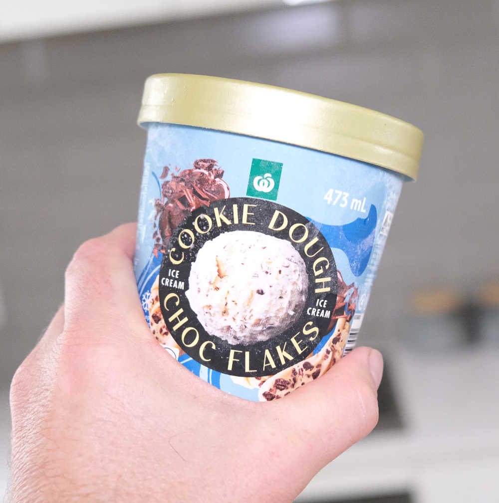 A hand holding a container of WOW Cookie Dough Choc Flakes Ice Cream with a blurred kitchen background.