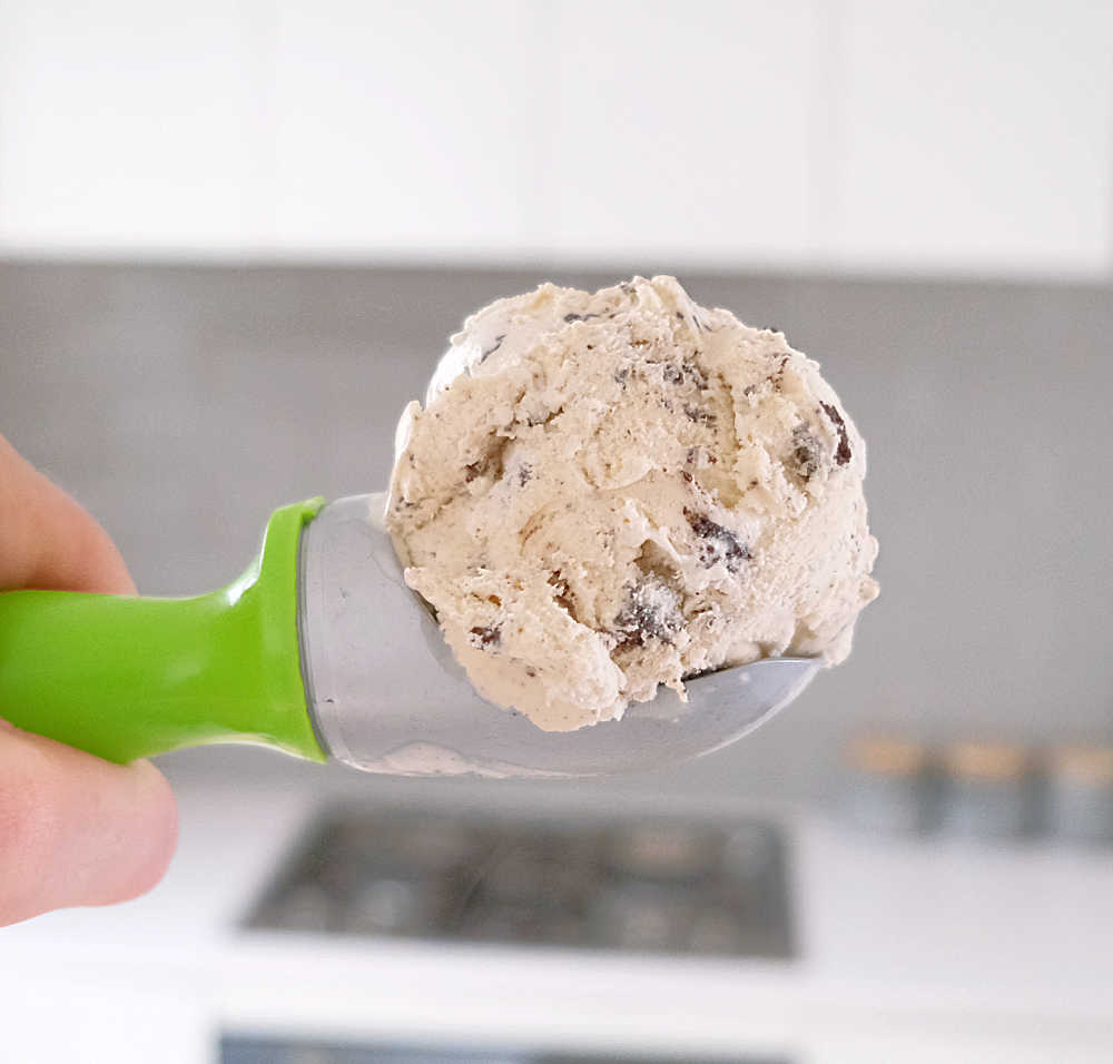 A hand holding a scoop of Bulla Cookies and Cream with a blurred kitchen in the background.