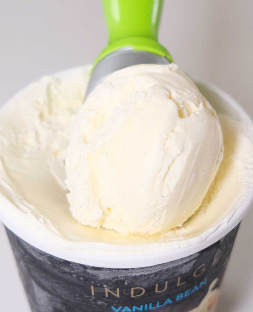 Closeup of a scoop of Indulge Vanilla Bean Ice Cream lying on the top of the opened tub.