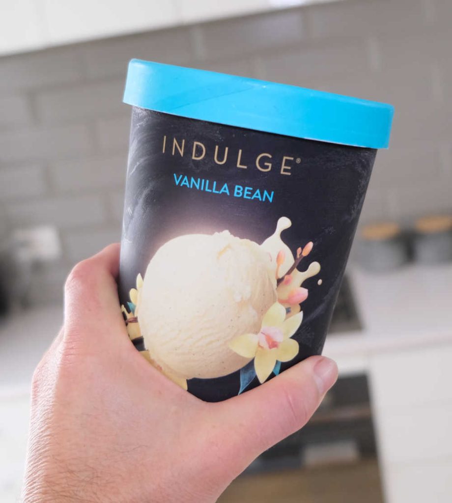 A hand holding a tub of Indulge Vanilla Bean Ice Cream with a blurred kitchen in the background.