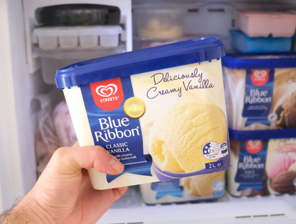 Holding a container of Streets Blue Ribbon Vanilla with a home freezer in the background.