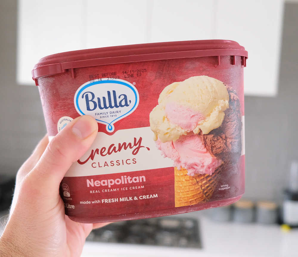 A hand holding a container of Bulla Neapolitan Ice Cream 2L.