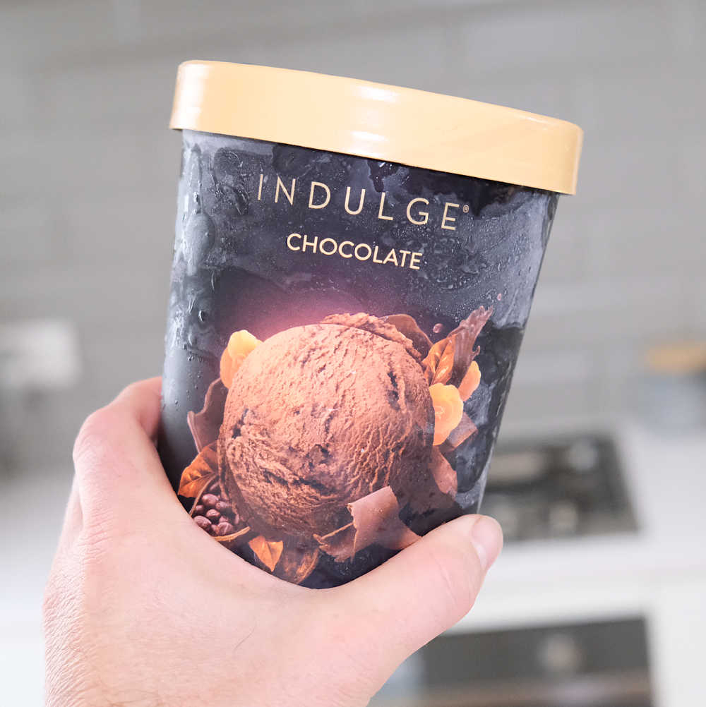 Holding a container of Indulge Chocolate Ice Cream 1L with a blurred kitchen in background.