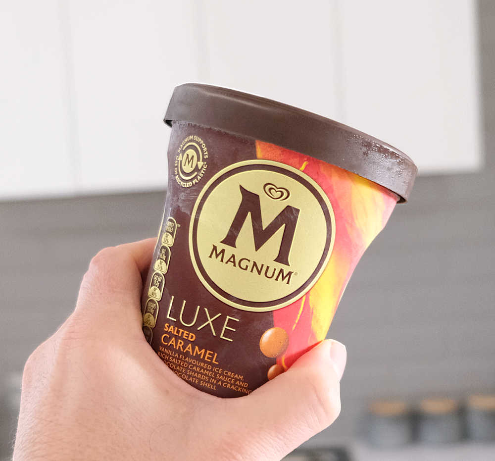 Holding a container of Magnum Luxe Salted Caramel with a blurred kitchen in the background.