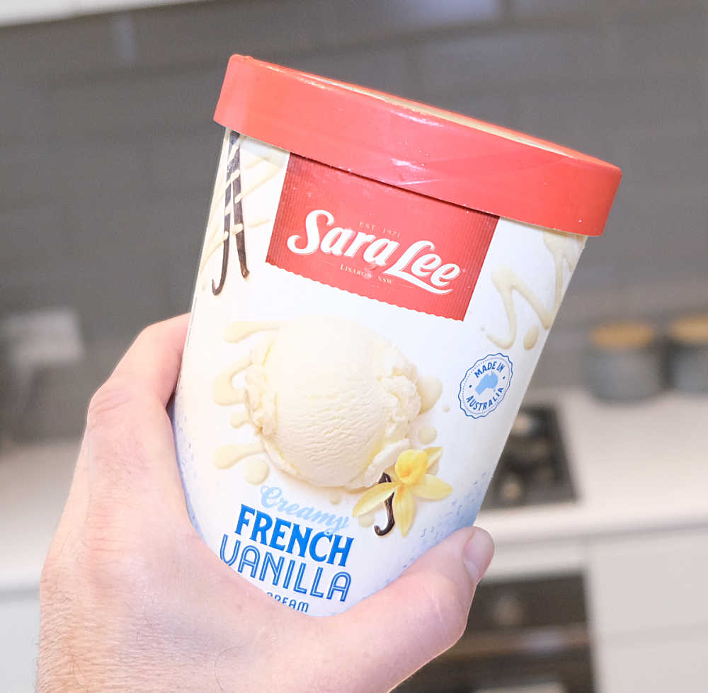 A hand holding a pack of Sara Lee French Vanilla Ice Cream with a blurred kitchen in the background.