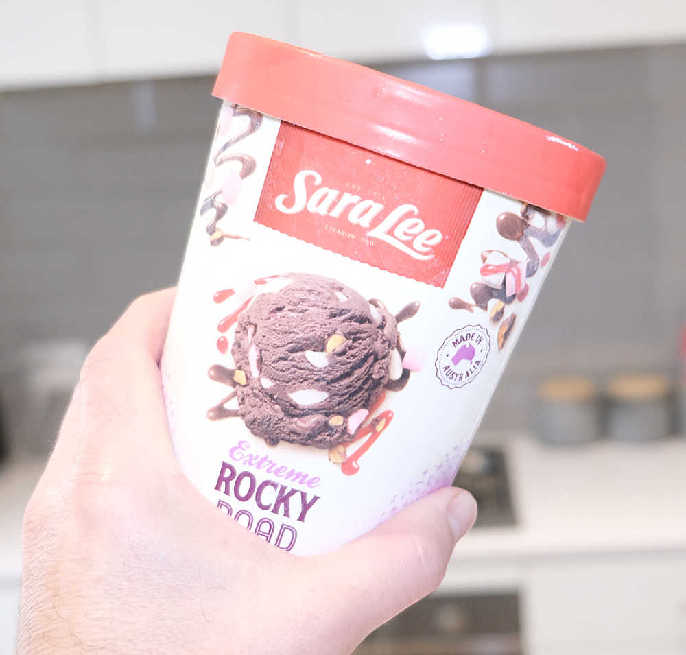 A hand holding a pack of Sara Lee Rocky Road Ice Cream with a blurred kitchen in the background.