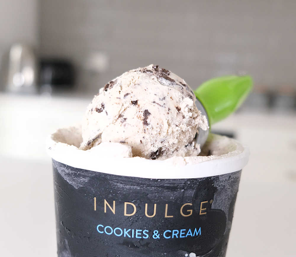 A scoop of Indulge Cookies and Cream Ice Cream resting on the top of the tub.