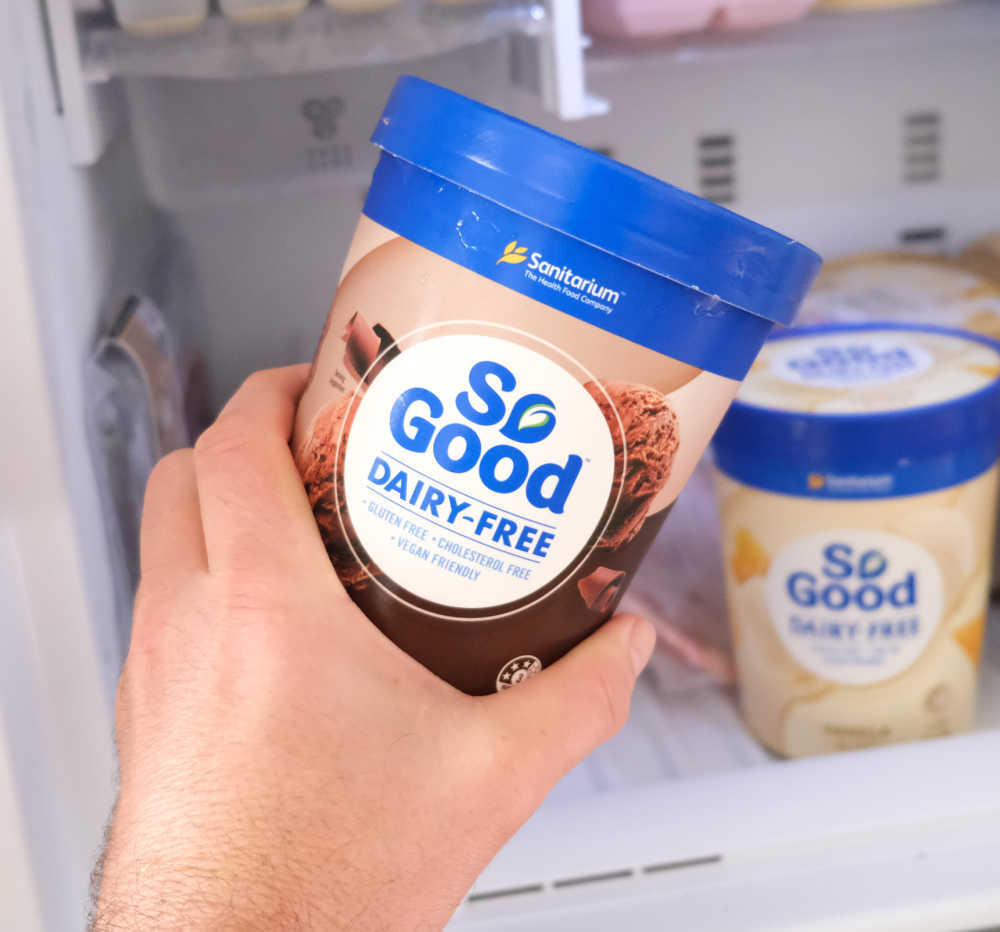 Holding a container of So Good Chocolate Bliss 1L with a freezer in the background.