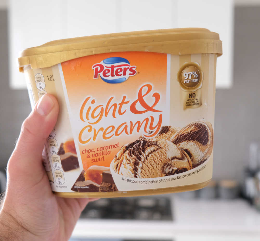 A hand holding a pack of Light and Creamy Choc, Caramel and Vanilla Swirl Ice Cream with a blurred kitchen in the background.
