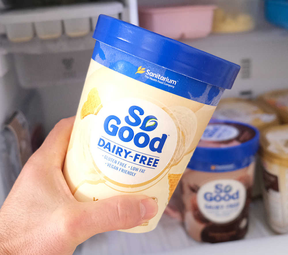 Holding a pack of So Good Vanilla Bliss 1L with a home freezer in the background.