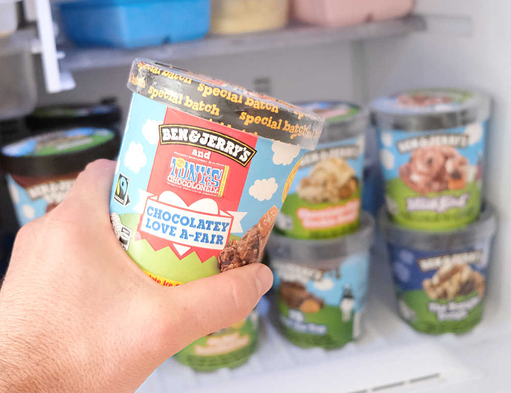 Holding a pint of Ben & Jerry's Chocolatey Love A-Fair with a home freezer in the background.