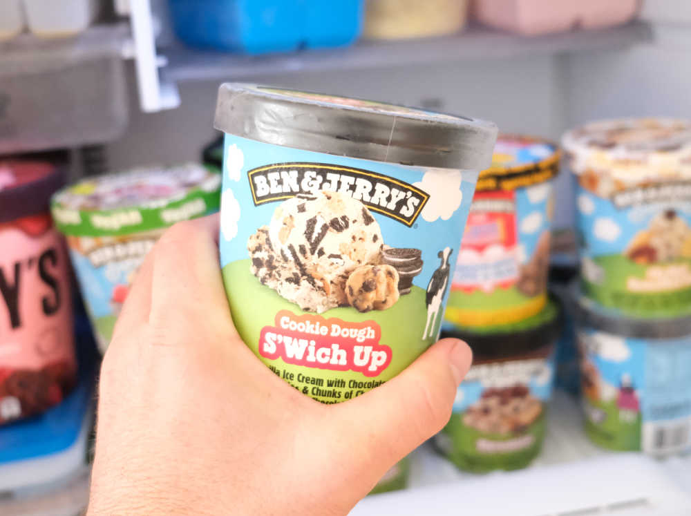 Holding a pack of Ben & Jerry's Cookie Dough S'Wich Up with a home freezer in the background.