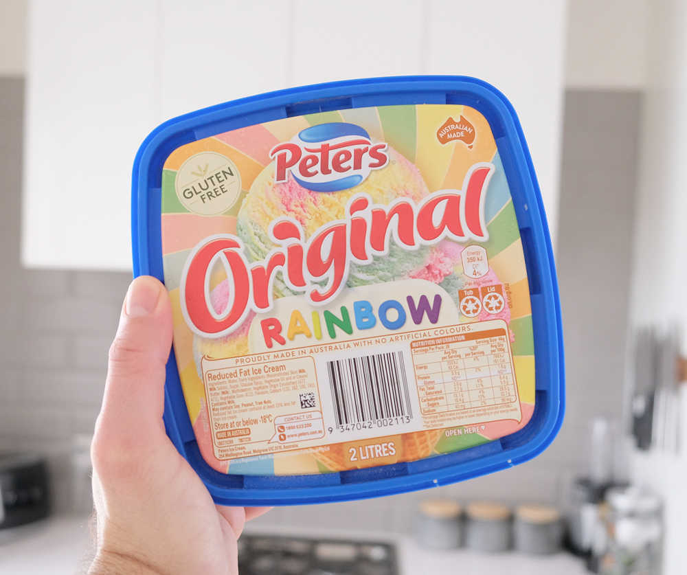 Holding a container of Peters Original Rainbow 2L with a kitchen in the background.
