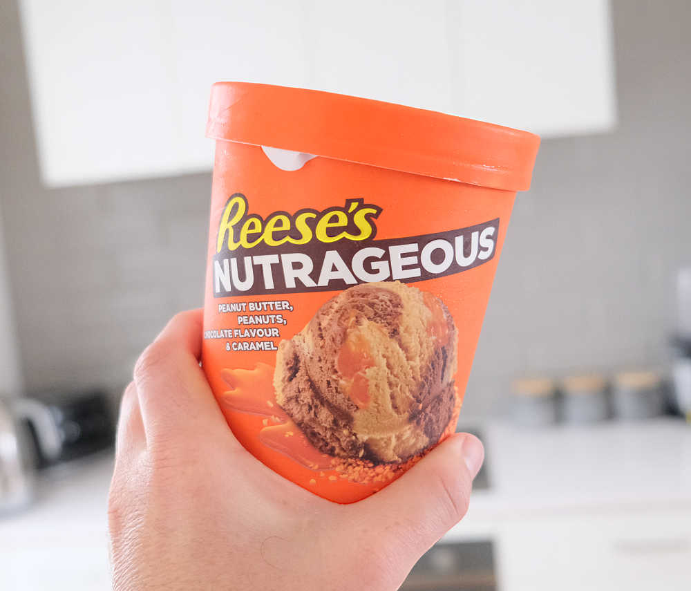 Holding a container of Reese's Nutrageous 1L with a kitchen in the background.