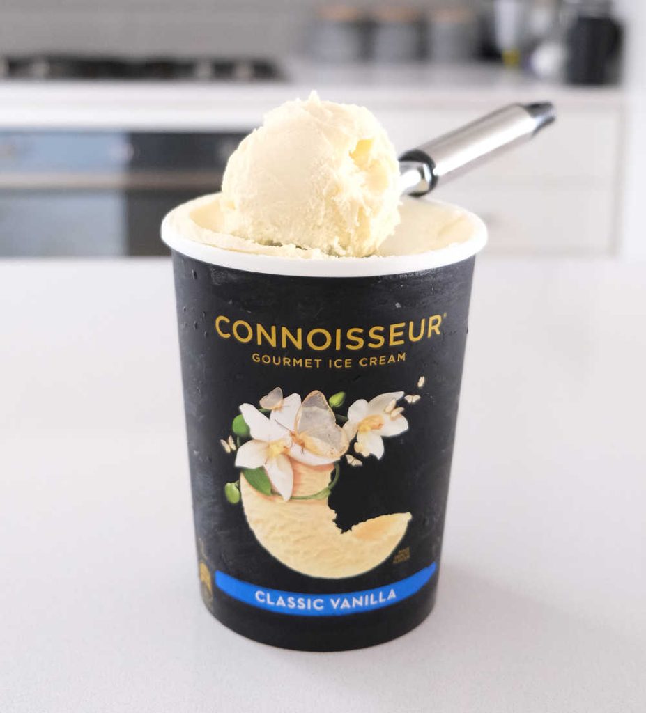 A pack of Connoisseur Gourmet Ice Cream Classic Vanilla on a kitchen bench with a scoop resting on the top of the pack.